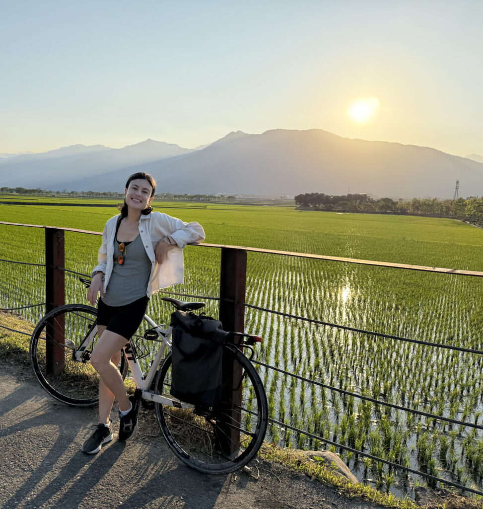 A photograph of Tara leaning against the fence of a bright green rice field, with a Taiwanese mountain range and sun setting in the background.
