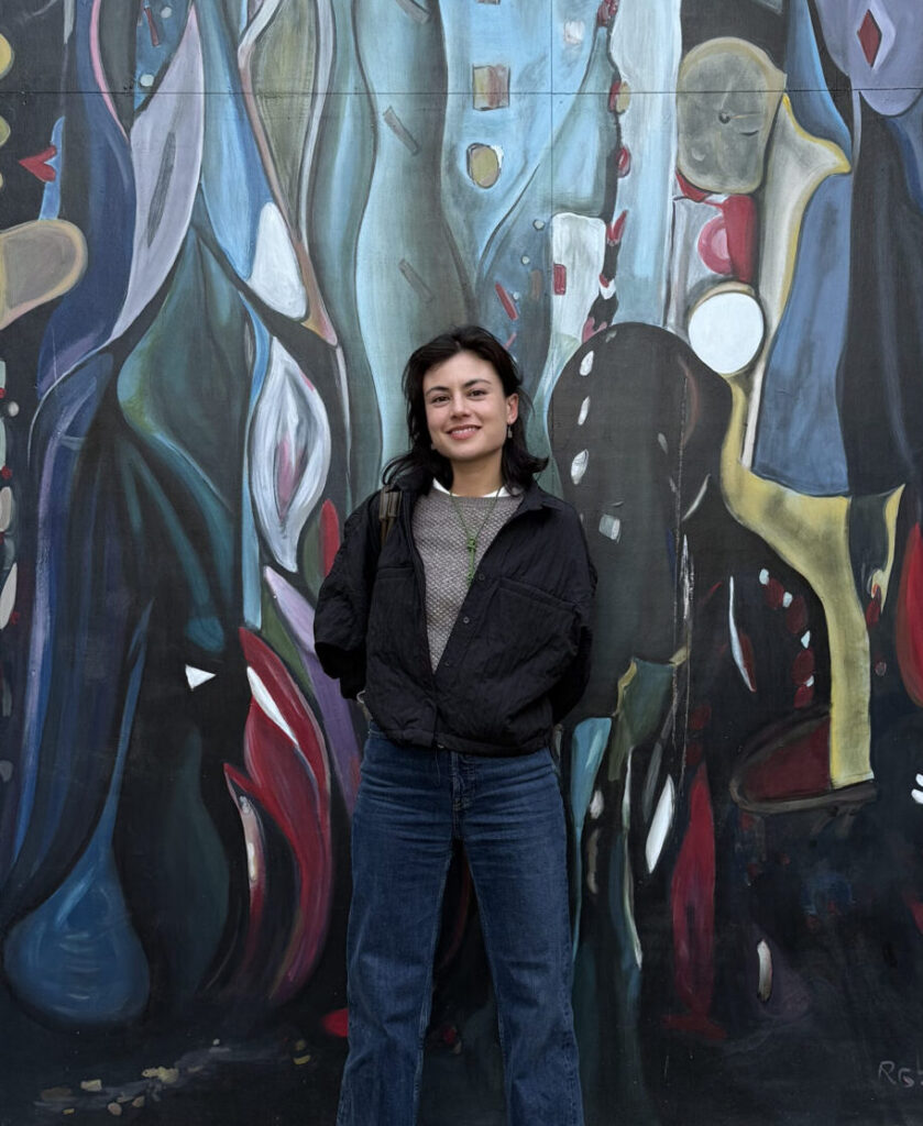 Tara standing in front of a large wall mural of abstract organic shapes and dark moody hues of blue, purple, green, red, and black.